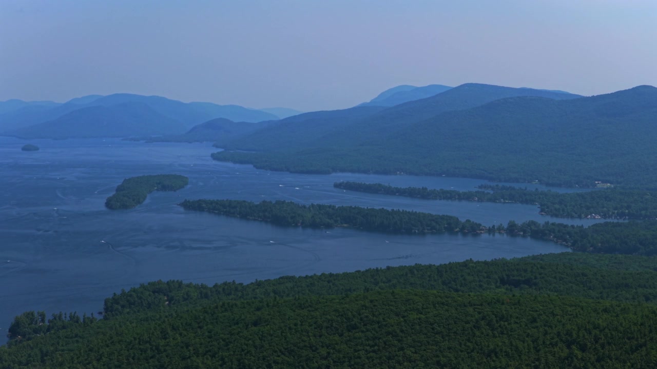 Punto di ritrovo di Lake George, nello stato di New York, in una soleggiata giornata estiva di luglio. Vista aerea con drone della città di Lake George, isola, Monti Adirondack, porto con barche e cielo blu mattutino, con movimento circolare a destra.