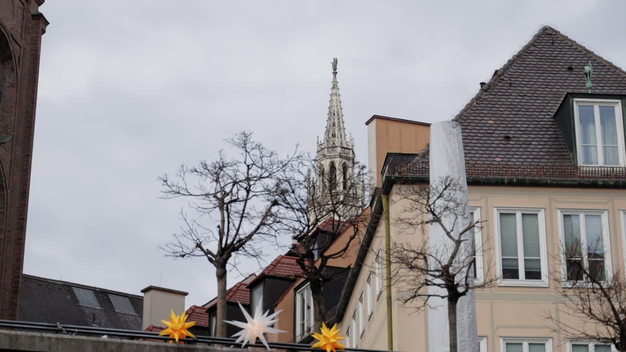 A Gothic church spire rises above Munich’s rooftops on a cloudy winter day. Bare trees and festive star decorations create a contrast between seasons, blending historical and modern elements.