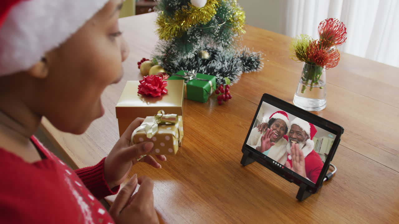 mujer afroamericana con sombrero de santa usando tableta para una videollamada de navidad con una pareja en la pantalla