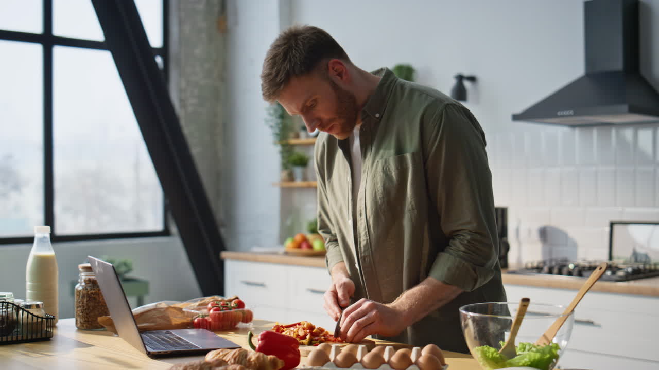 Serious man cutting vegetables in kitchen watching online recipe laptop closeup