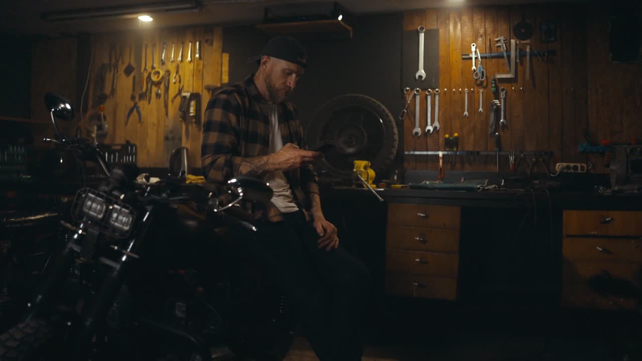 A mechanic guy sits on his moped and watches social networks on his phone during a break at work in the workshop