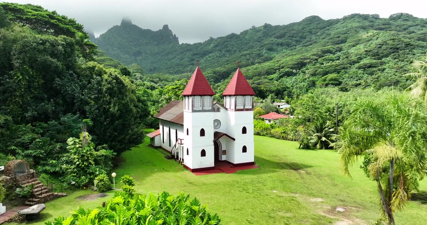 Le due guglie con tetto rosso della chiesa di Lisa si ergono sopra un prato curato incorniciato da palme, con una cima vulcanica frastagliata che incombe dietro una fitta valle di giungla tropicale nella Polinesia Francese. Vista aerea con drone