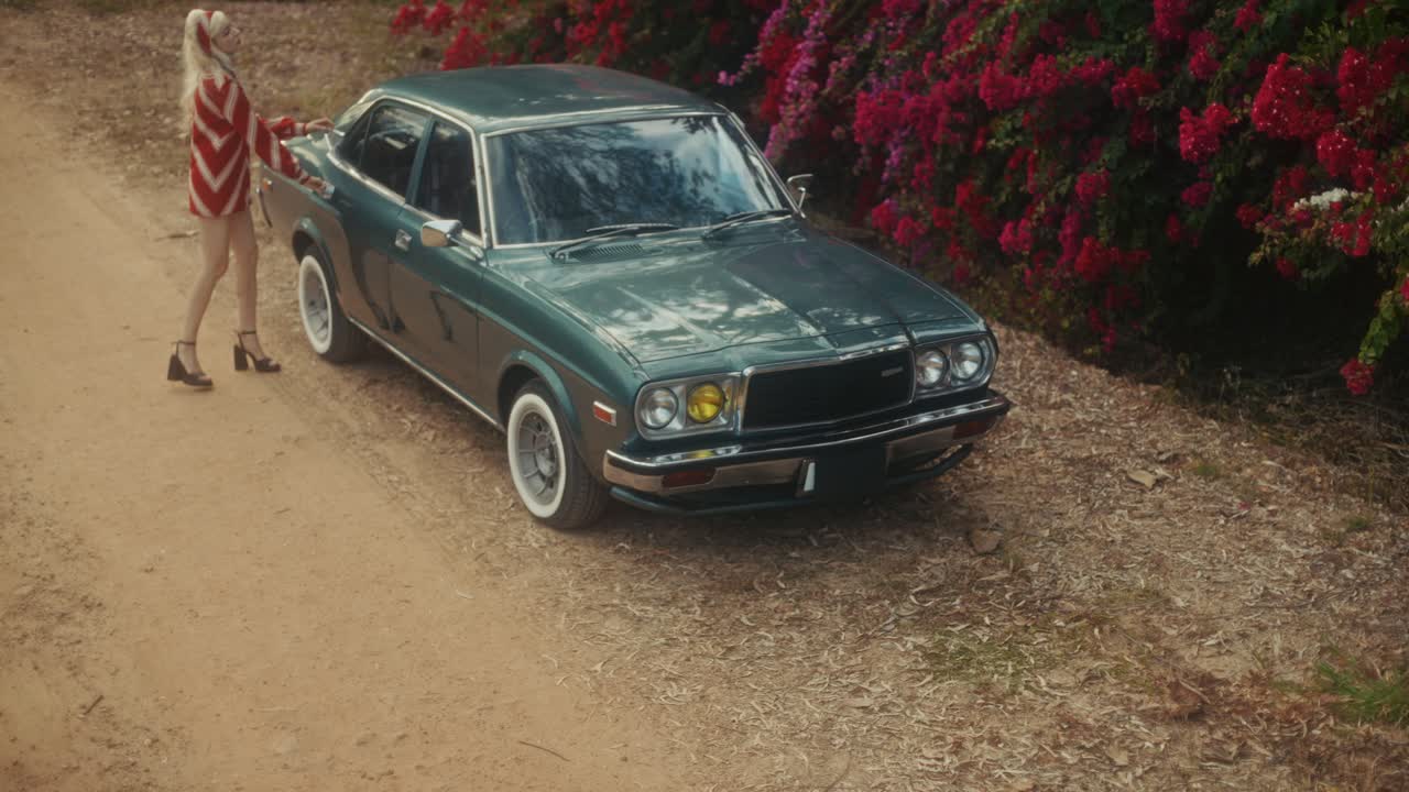 Mujer elegante saliendo de un coche antiguo con una maleta roja en un camino de tierra