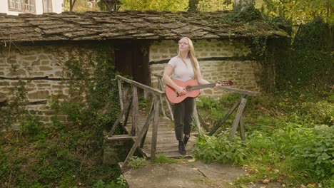 Beautiful-young-woman-strums-acoustic-guitar-rustic-wooden-bridge