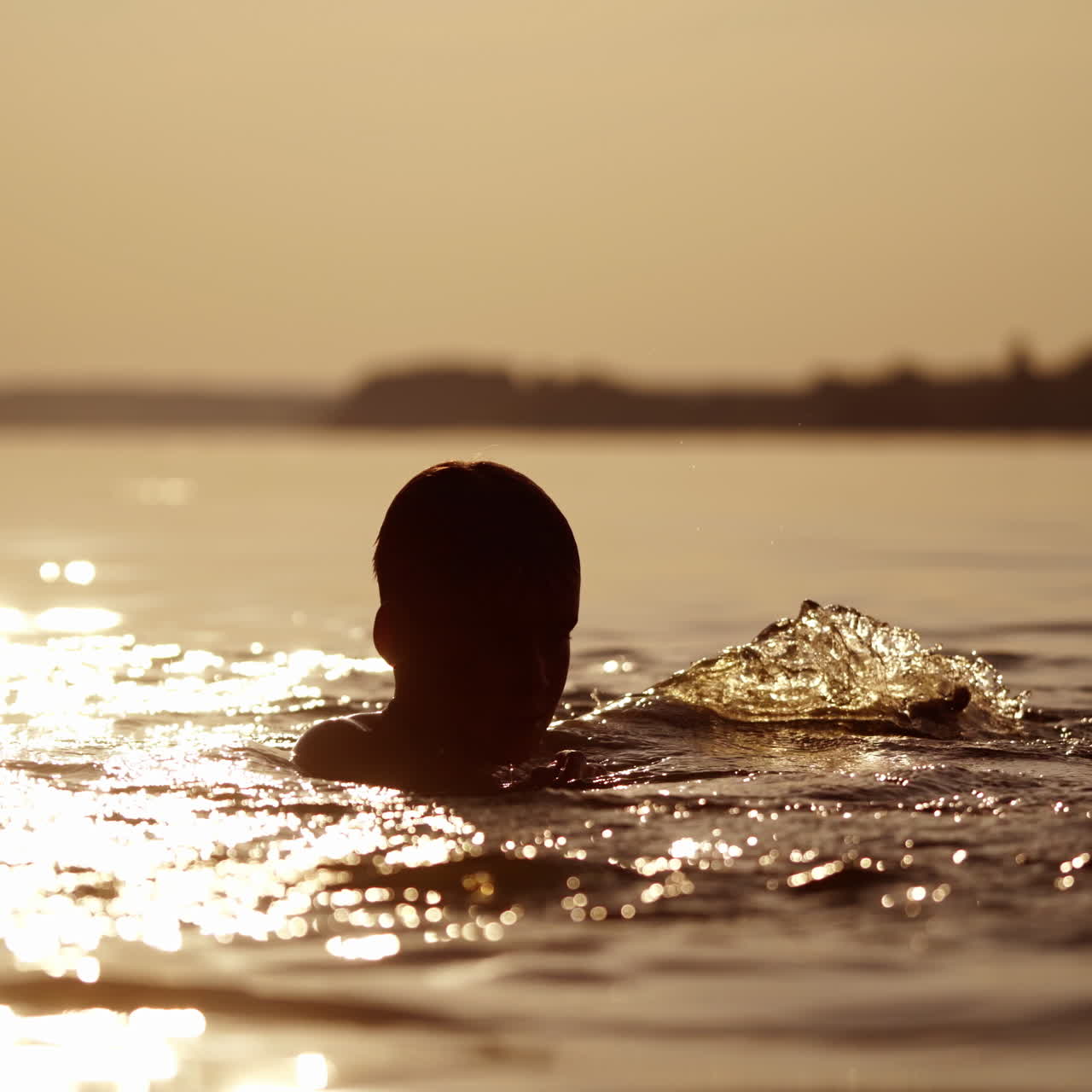 Young boy plays with water in the evening river. Silhouette of a child making waves on natural water background at sunset. Healthy lifestyle.