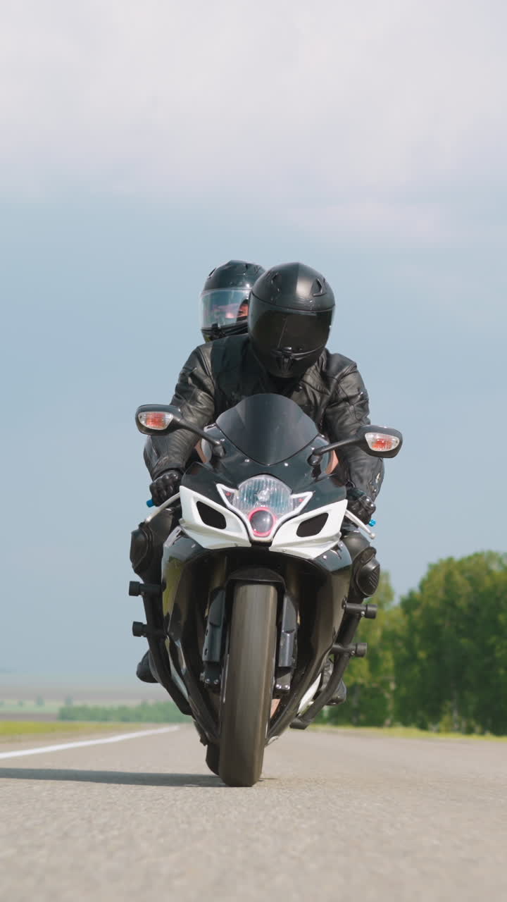 Skilled biker with passenger rides powerful motorcycle along asphalt road past protective forest belt under sky with clouds at countryside slow motion