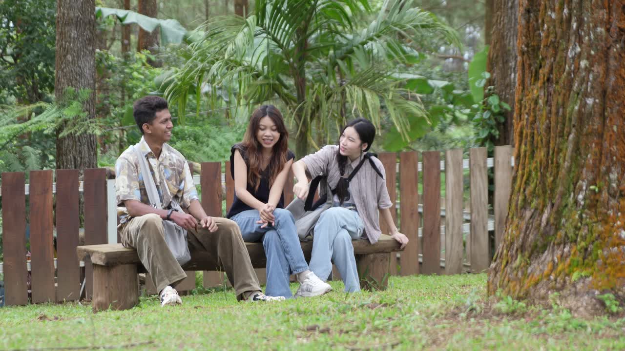 เพื่อนชาวเอเชียอินโดนีเซียวัยหนุ่มสาวกำลังนั่งเล่นด้วยกันบนม้านั่งไม้ในสวนป่า
