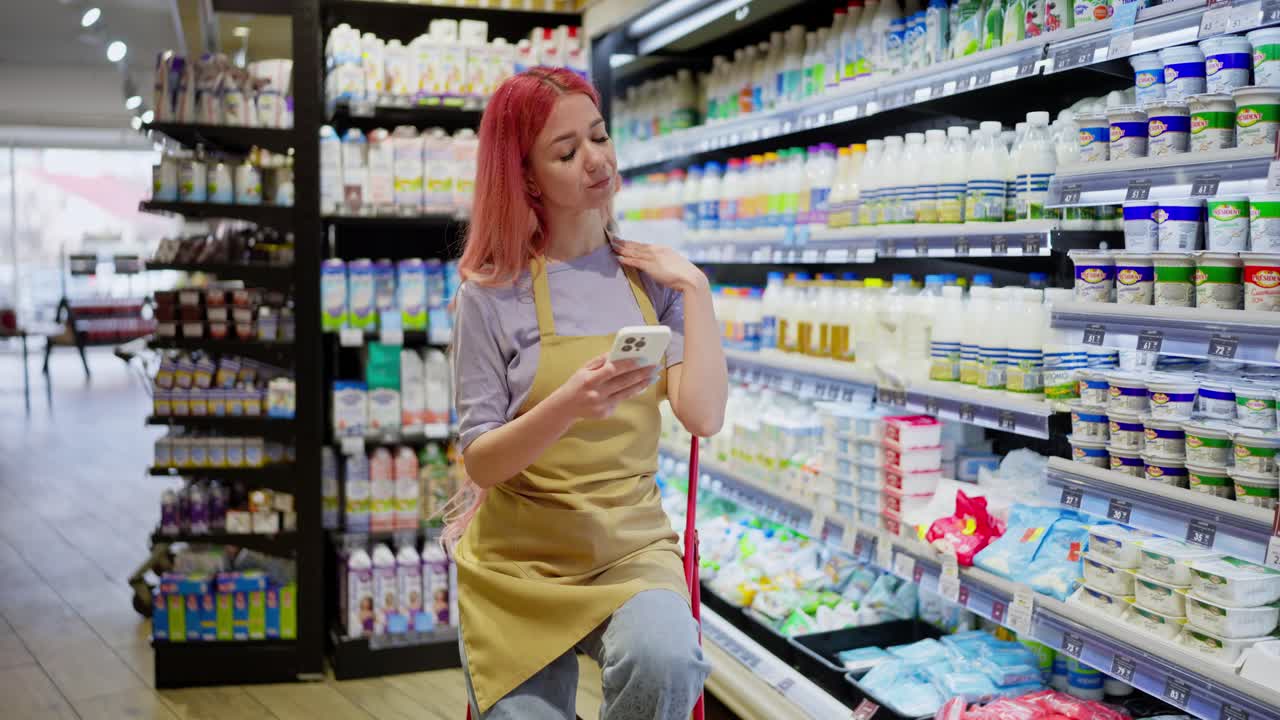 분홍색 머리카락을 가진 피곤한 소녀는 휴식을 취하고 슈퍼마켓의 유제품 부서에서 일하면서 드폰으로 소셜 네트워크를 스크롤합니다.