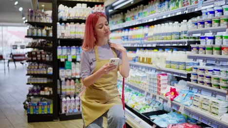 una chica cansada con el cabello rosa toma un descanso y se desplaza a través de las redes sociales en su teléfono mientras trabaja en el departamento de lácteos de un supermercado