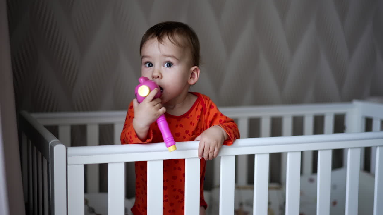 Female hand gives a toy to a baby standing in the crib. Sweet little baby takes a toy and shoves it to his mouth looking up. Close up.