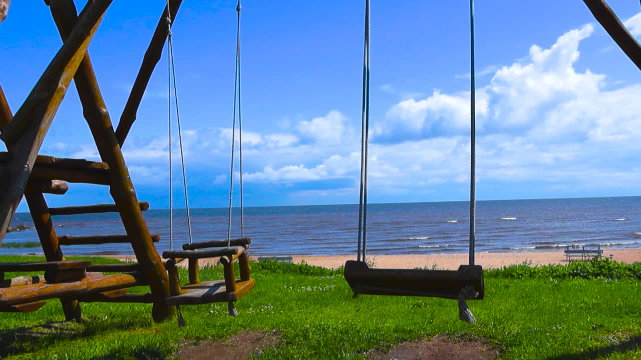 Static back view in slow motion empty swing gently swaying in front of sunlit beach, Peipsi Lake. Wooden swingset play structure. Blue sky with white clouds, waves roll toward the shore in background