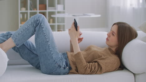 Frau-Liegt-Auf-Der-Couch-Im-Wohnzimmer-Und-Chattet-Mit-Dem-Smartphone,-Um-Eine-Nachricht-Zu-Schreiben.-Einsame,-Glückliche-Dame,-Die-Sich-Mit-Ihrem-Smartphone-Entspannt,-Auf-Der-Couch-Sitzt-Und-Liest,-Schreibt,-Sucht-Und-Im-Mobilen-Internet-Surft