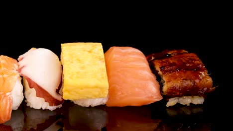 a neatly arranged selection of sushi nigiri and sashimi, including shrimp, salmon, tuna, egg, and roe, displayed on a glossy black surface under bright studio lighting with a slow left-to-right camera pan