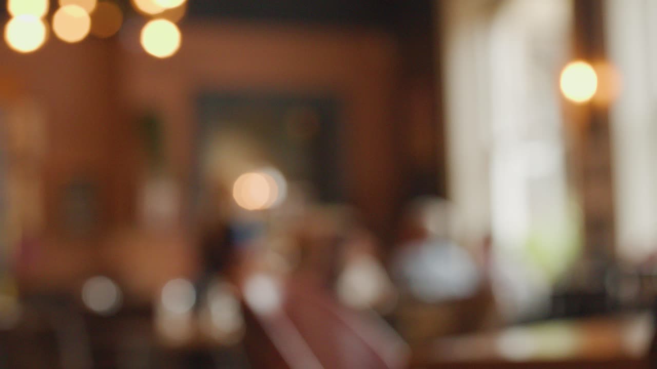 Soft-focus view of a warmly lit restaurant or pub interior, with indistinct figures and tables. Ambient lighting, shallow depth of field, and static camera create a relaxed, inviting mood