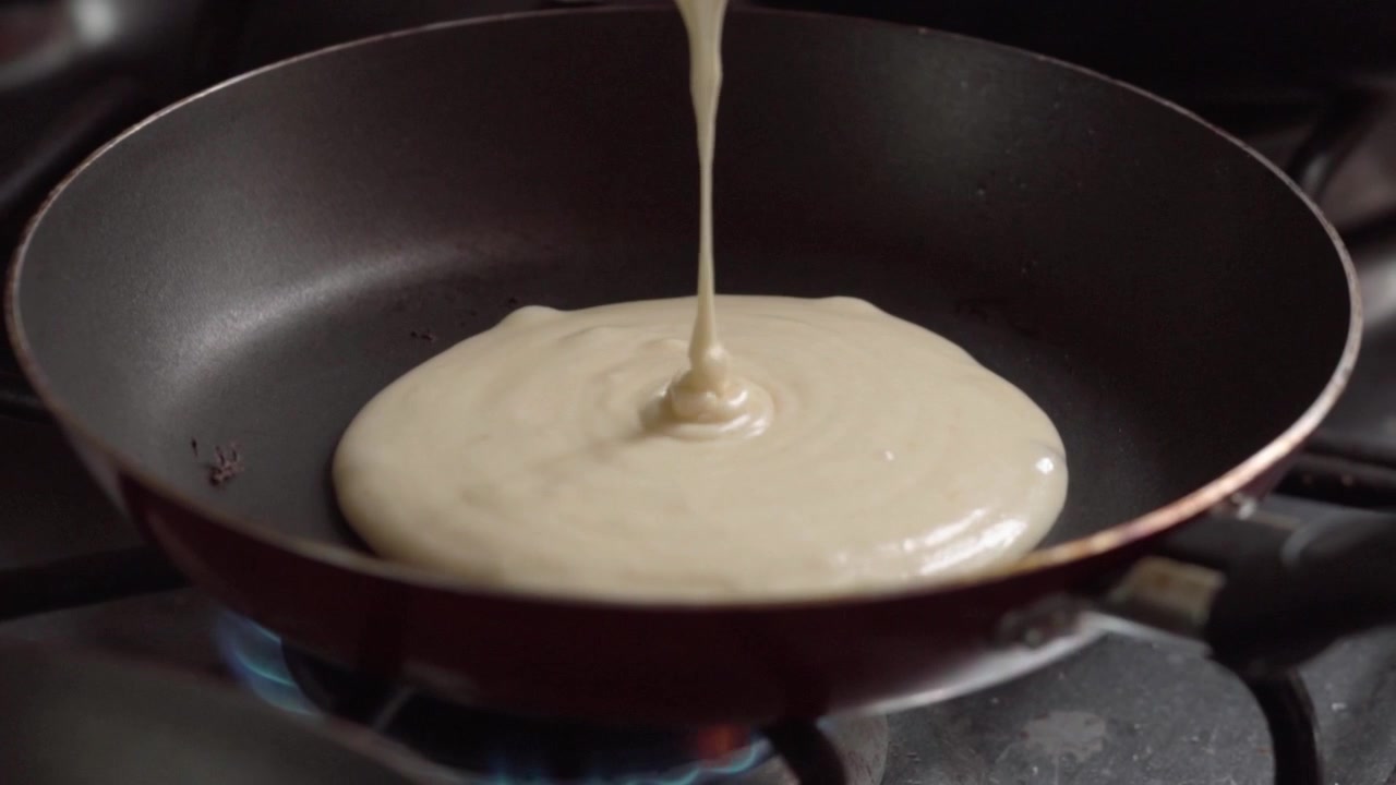 Fresh pancake batter pouring into hot frying pan over gas flame during breakfast preparation. Home cooking process showing smooth mixture spreading before flipping stage