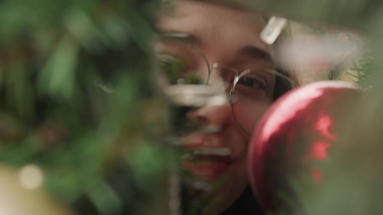 Close up white woman student peeking through evergreen branches at red bauble, wearing round glasses, soft bokeh and warm natural light, intimate holiday moment with curious expression