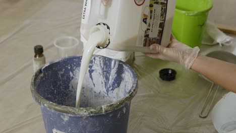 person wearing gloves pouring white paint from a large can into a blue bucket