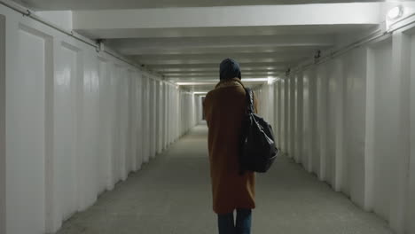 A back shot of a girl wearing a brown coat, jeans, white shoes, and a backpack as she slowly walks through an underpass or tunnel. The scene captures a sense of solitude and contemplation