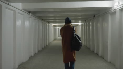 a back shot of a girl wearing a brown coat, jeans, white shoes, and a backpack as she slowly walks through an underpass or tunnel. the scene captures a sense of solitude and contemplation