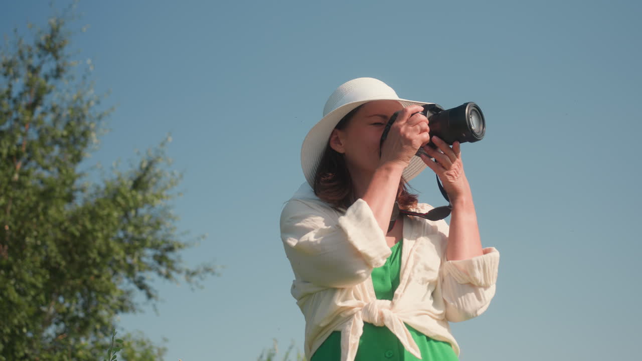 Beautiful woman in green dress and white hat looks at camera in hand while descending sunny grassy hill surrounded by summer flora, preparing to take nature photo under clear blue sky