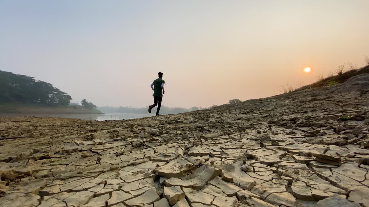 toma en ángulo bajo de un corredor corriendo en el lecho del río seco, suelo agrietado, cielo brumoso