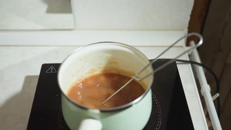 Food steaming on electric stove in light green pot with whisk resting inside, mixture bubbling with thick consistency under heat, countertop and reflection in mirror behind