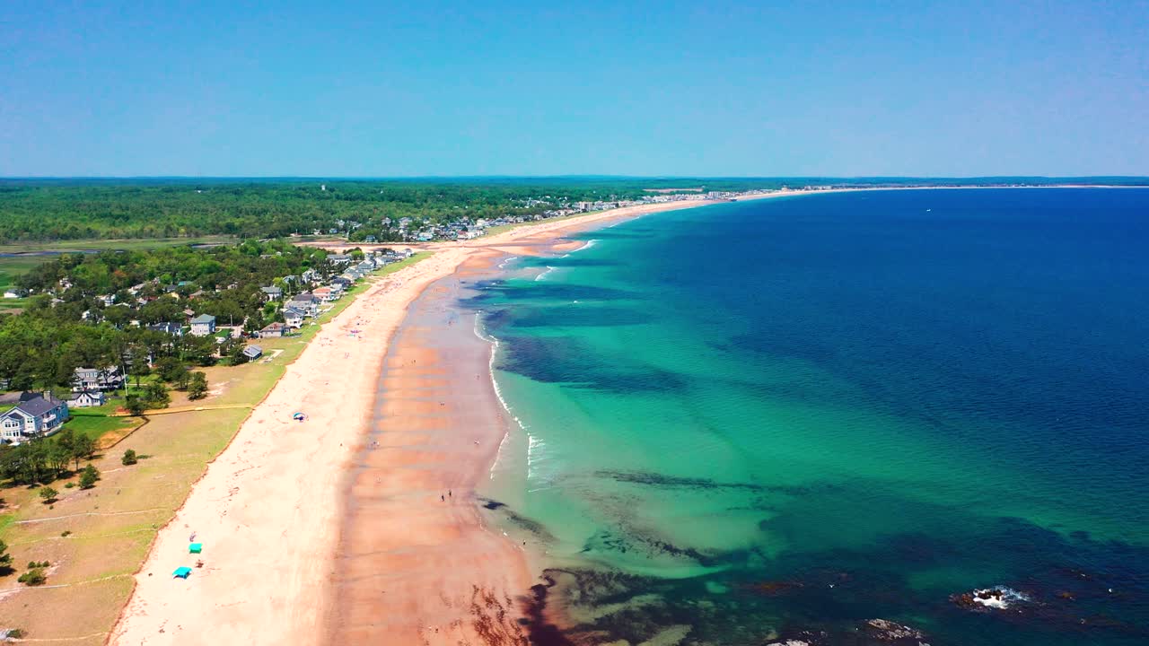 Drone captures vibrant beaches in Saco, Maine. Tourists walk, play, and relax on sunny sand as Atlantic waves roll in. Vacation homes line the coastline, showcasing New England’s summer seashore charm