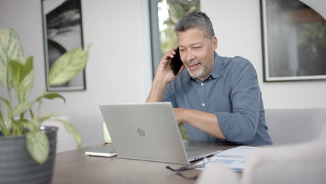 un hombre bi-racial feliz hablando en su teléfono inteligente y usando una computadora portátil, trabajando desde casa, en cámara lenta.