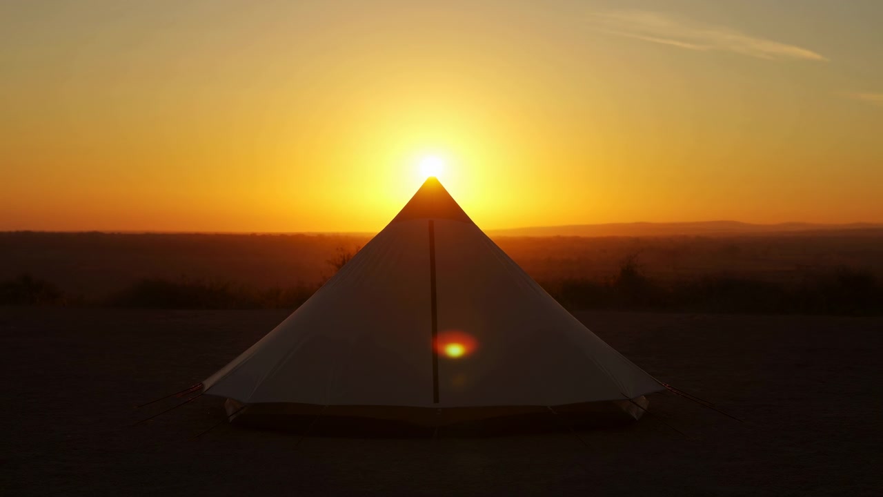 Tenda triangolare bianca si erge centrata su terreno aperto al tramonto, con il sole splendente allineato sopra la cima, colline distanti all'orizzonte, cielo ambrato caldo, leggera foschia, bagliore sottile e un'atmosfera calma da campeggio remoto