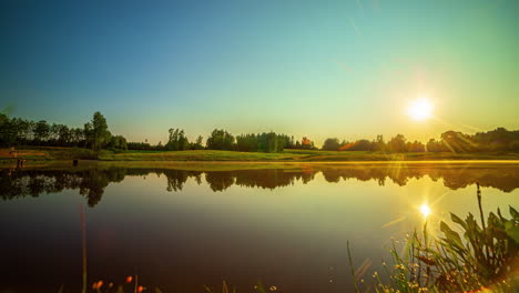 Hermoso-Lapso-De-Tiempo-De-Una-Puesta-De-Sol-Sobre-Un-Lago-Y-Un-Campo,-Mientras-El-Sol-Se-Refleja-En-El-Lago-Y-La-Niebla-Vespertina-Se-Pone