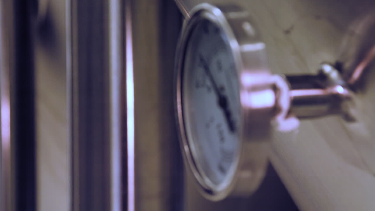 Rack focus between a close-up of a temperature gauge mounted on a stainless steel fermentation tank in a brewery to background and back
