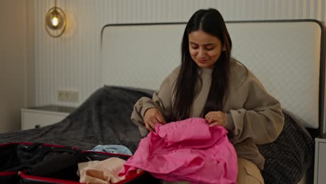 a happy and confident brunette girl with brown skin in a gray sweater puts her pink blue and beige things in a black suitcase while preparing for the weekend and leaving travel in a modern apartment in the bedroom