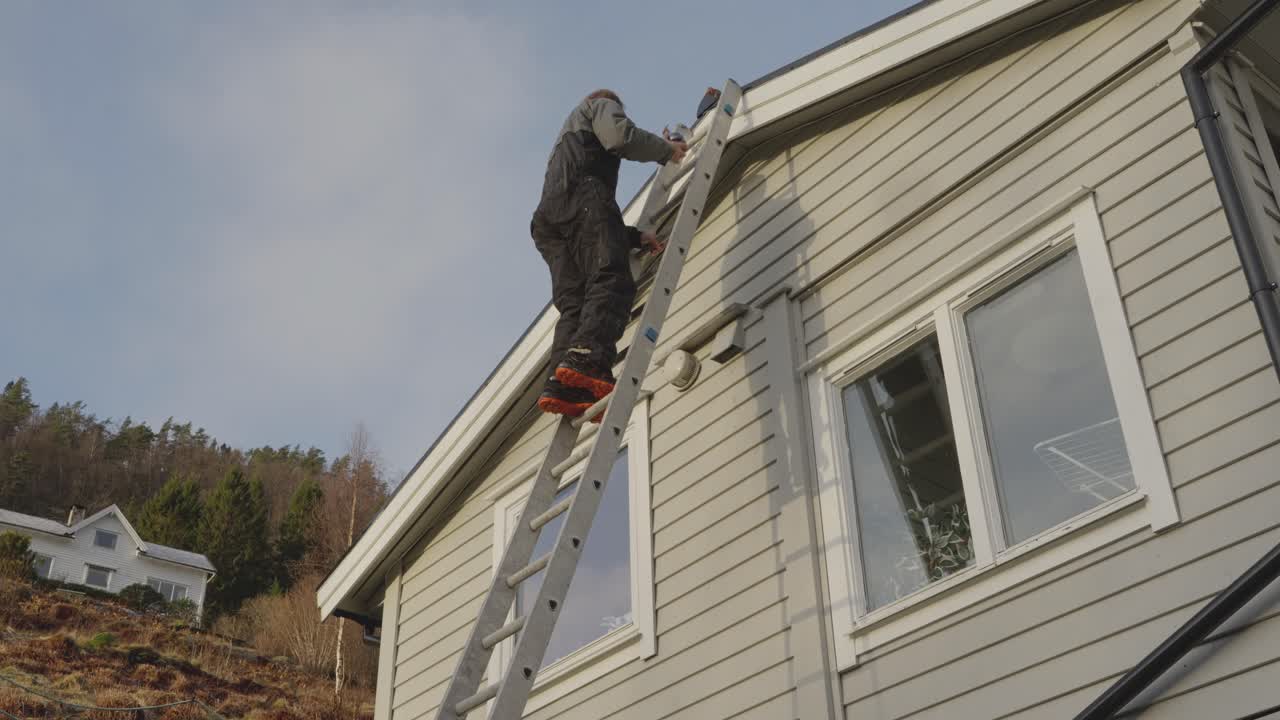 Person climbs ladder to mount a black security camera under the roof of a private house, completing installation and removing the ladder