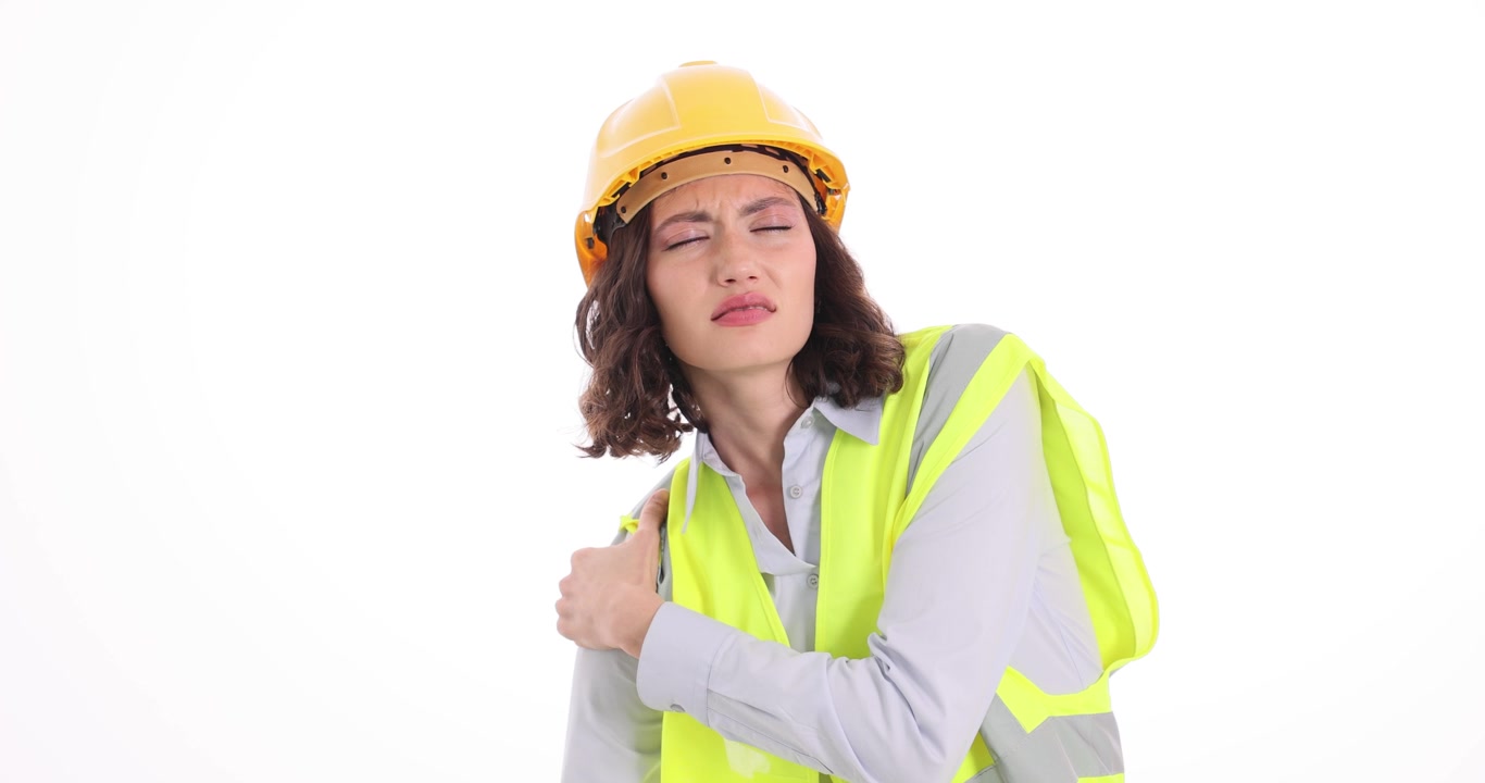 Trabajadora con chaleco de seguridad y casco se masajea el hombro dolorido.