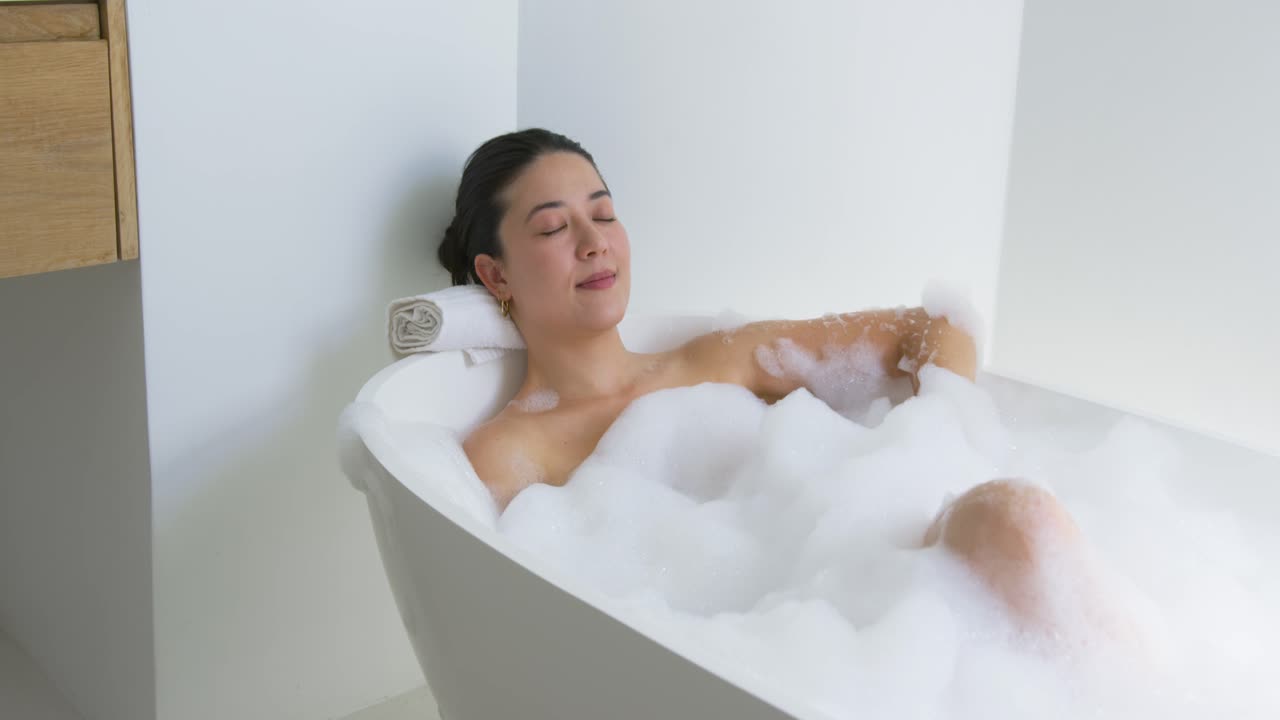 African American adult woman reclining in bathtub with foam, rolled towel headrest, opening eyes