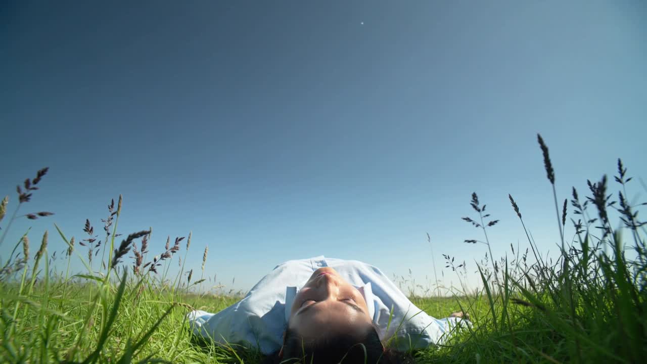 Mujer relajándose y respirando lentamente en un prado verde, con camisa de color claro, el sol calienta su piel.