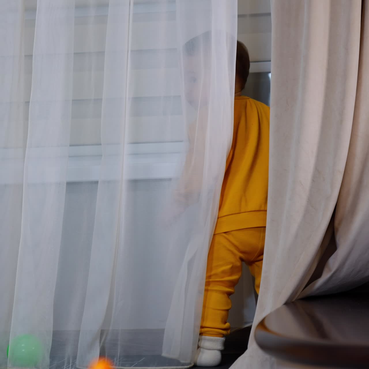Little kid standing at the window and hiding behind the curtain. Child playing indoors at the window-sill