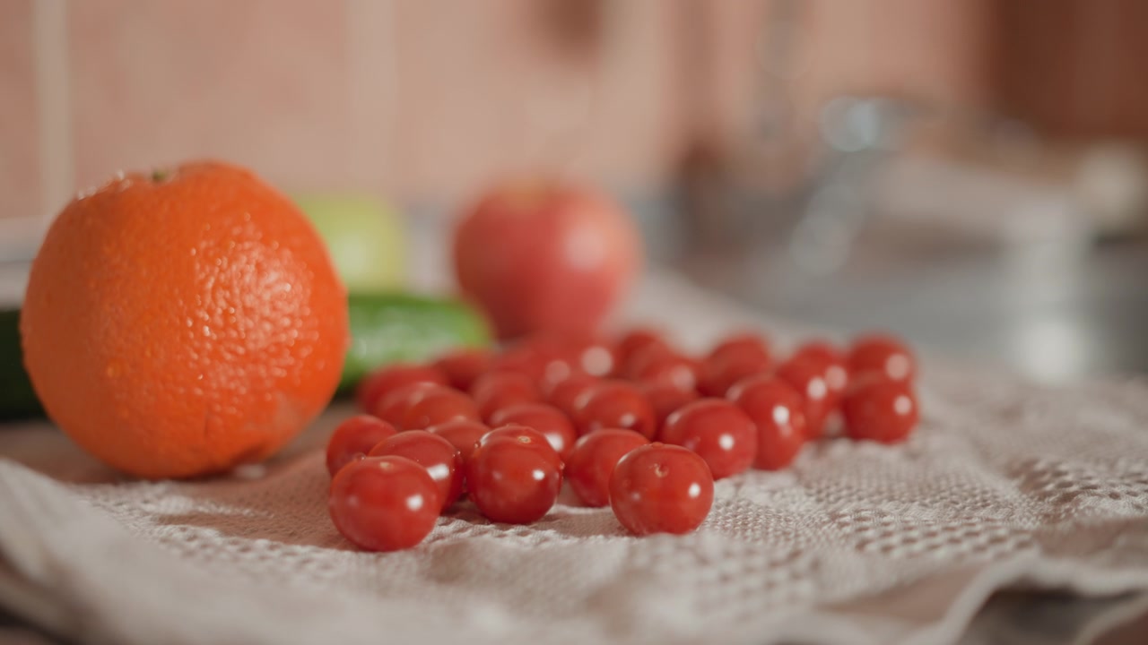 Lav vinkel af blanke cherrytomater arrangeret på tværs af et tekstureret køkkenhåndklæde ved siden af slørede appelsiner og blandede råvarer, med duggede overflader, blødt sidelys, varme toner og naturlig madstyling.