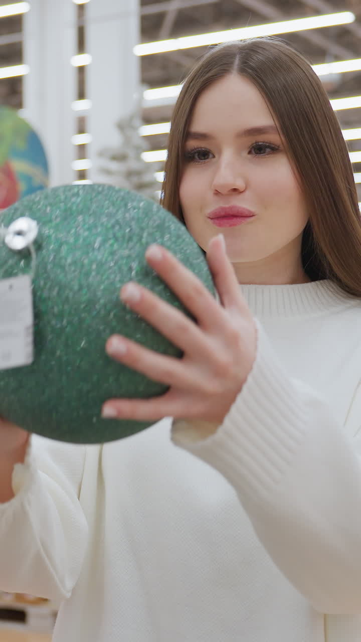 Young lady in a white sweater holding a large green shimmering Christmas ball ornament in a brightly lit decor store, surrounded by Christmas trees and festive decorations