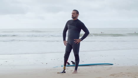 long shot of a confident bearded surfer in wetsuit with bionic leg standing on seashore, showing shaka sign and smiling at the camera