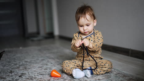 active caucasian baby boy sitting on the floor plays with a black cord. domestic black cat is watching the beautiful kid's play.