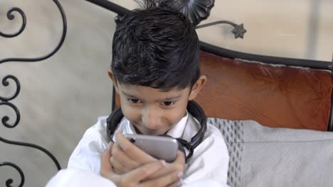 Lindo-Pequeño-Niño-Indio-Asiático-Caucásico-Mirando-Seriamente-La-Pantalla-Del-Teléfono-Móvil-Usando-Auriculares-En-El-Cuello-En-La-Sala-De-Estar-Vista-Frontal-De-Primer-Plano