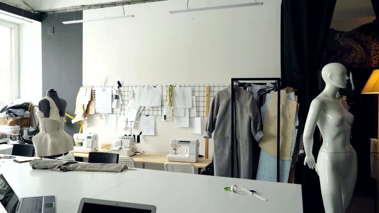 Pan shot of light clothing design studio with large tailor's desk, mannequins, numerous sketches pinned on wall, sewing machines and half-finished garments on rails.