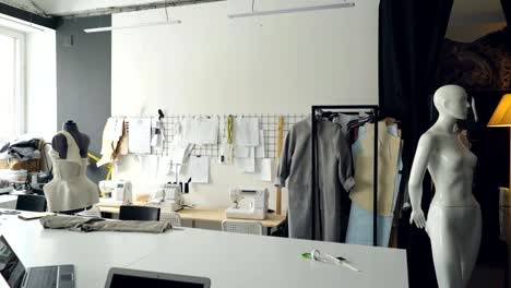 pan shot of light clothing design studio with large tailor's desk, mannequins, numerous sketches pinned on wall, sewing machines and half-finished garments on rails.