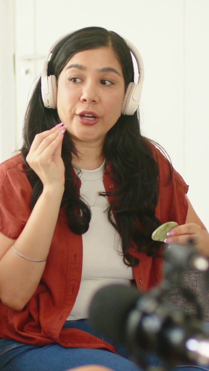 Video vertikal: Wanita memperbaiki headphone sambil menggunakan gua sha di pipi dan berbicara ke mikrofon di rumah, mengenakan atasan warna karat.