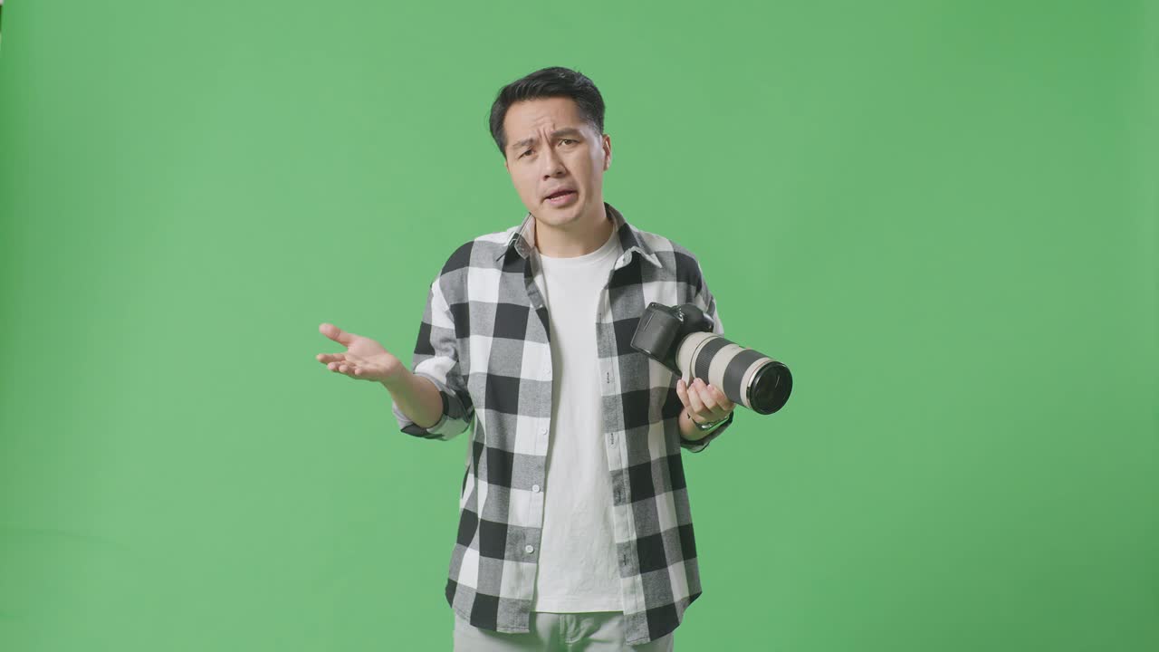 혼란스러운 아시아 사진작가는 손에 카메라를 들고, 왜라고 말하고 스튜디오에서 녹색 화면 배경에 서서 의심스럽게 몸짓을 합니다.