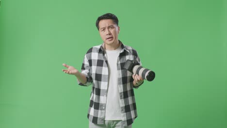 혼란스러운 아시아 사진작가는 손에 카메라를 들고, 왜라고 말하고 스튜디오에서 녹색 화면 배경에 서서 의심스럽게 몸짓을 합니다.