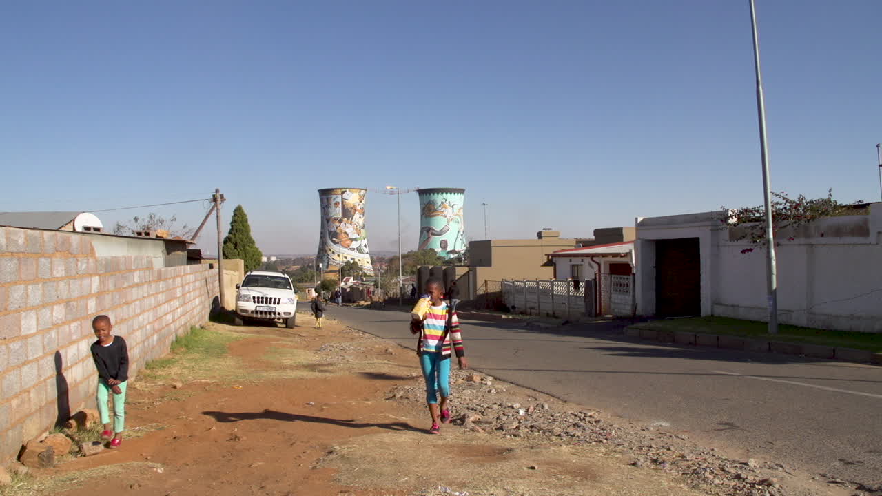 Young children in Soweto walk up a township street with the iconic Soweto cooling towers in the background.