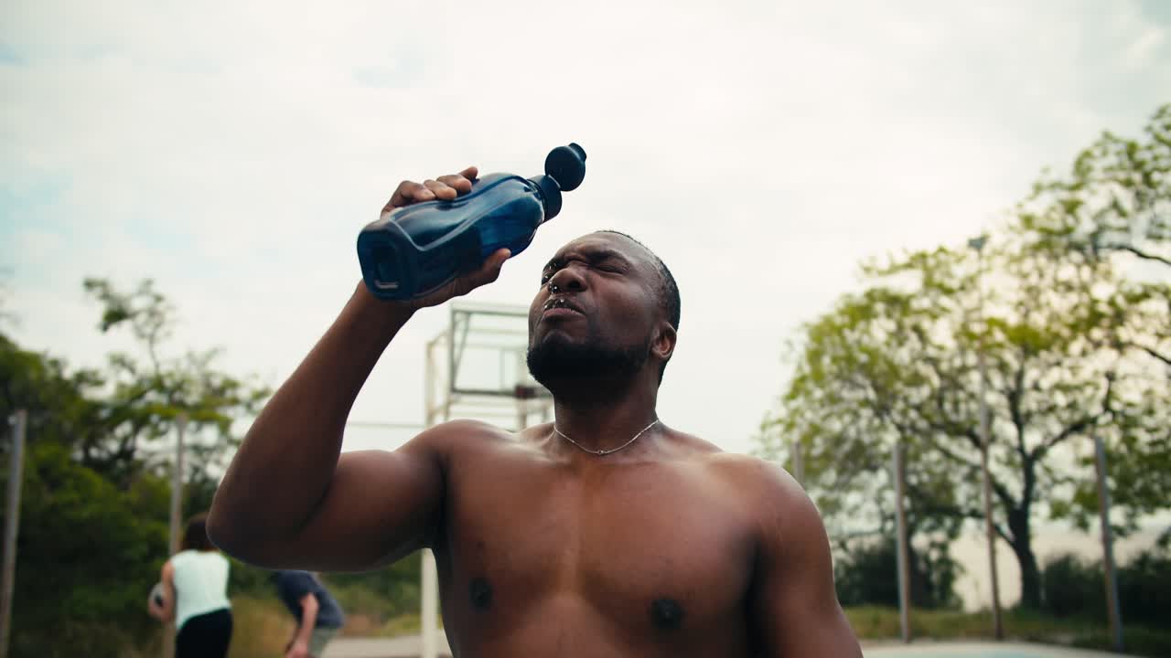 un hombre con piel negra y torso desnudo bebe agua en una cancha de baloncesto y luego lame su cabeza para refrescarse y grita su grito de batalla frente a sus amigos jugando baloncesto