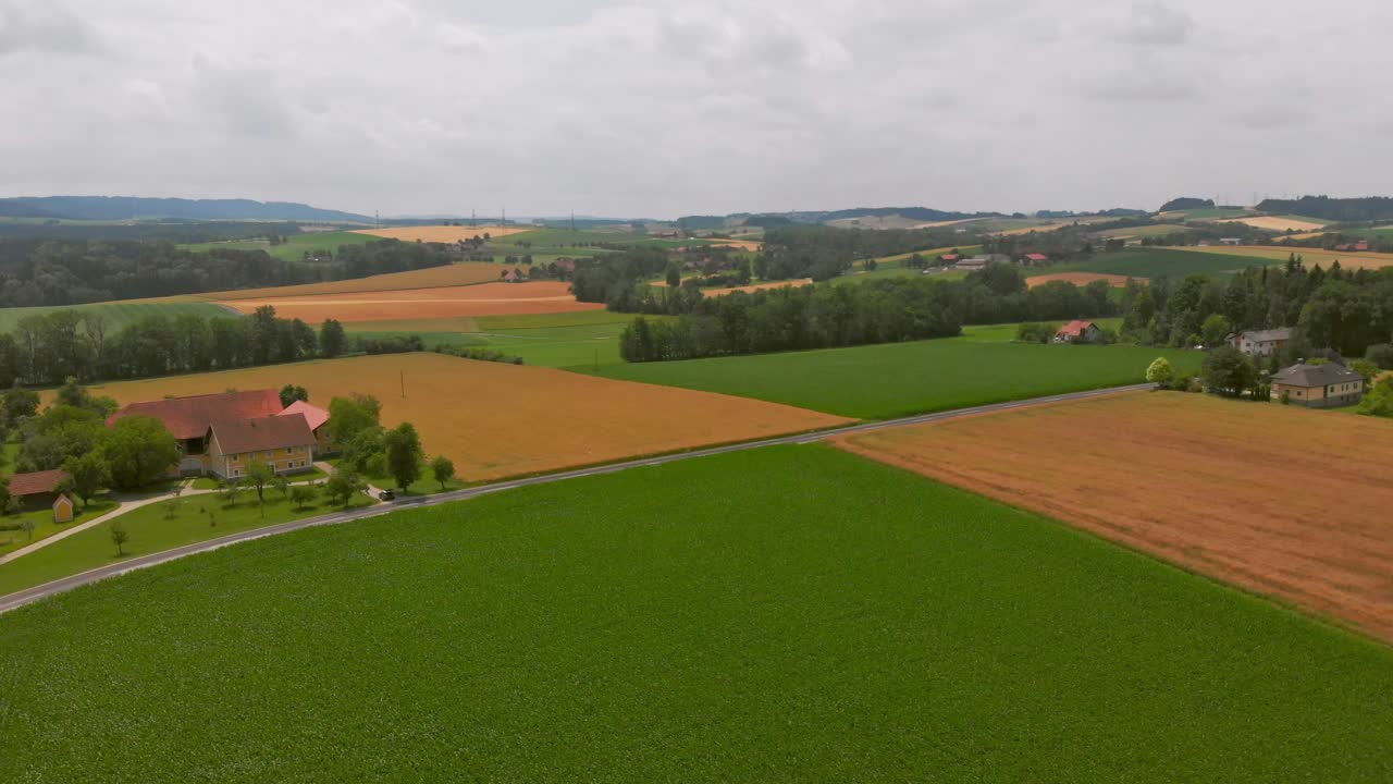 Aerial wide shot of rural farmland with patchwork fields, country road, scattered houses, and rolling hills under an overcast sky